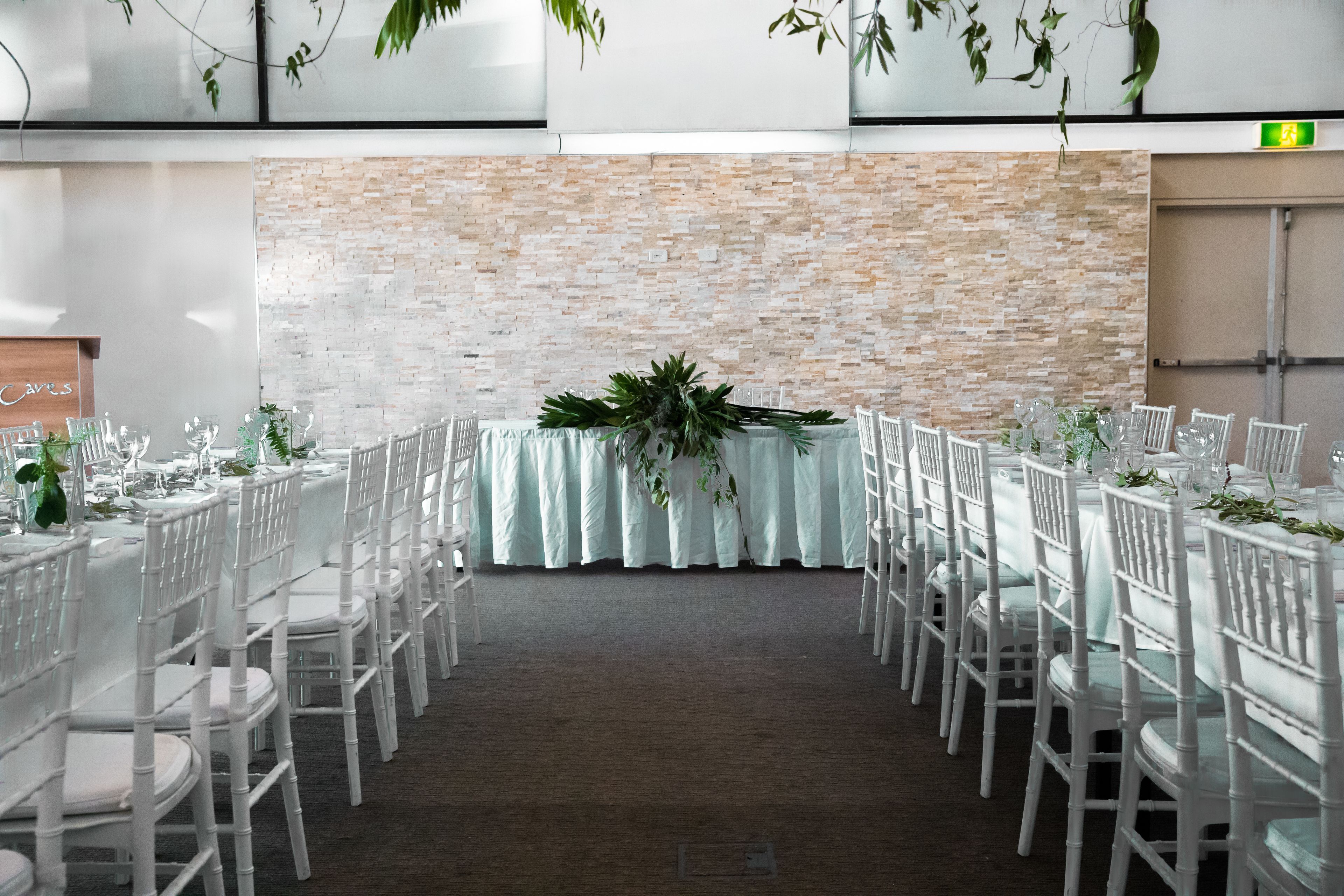 A banquet hall with white tables and chairs, a brick wall backdrop, and a central floral arrangement.