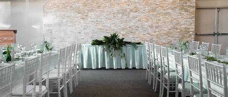 A banquet hall with white tables and chairs, a brick wall backdrop, and a central floral arrangement.