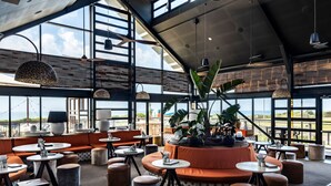 Un vaste espace extérieur aménagé avec des tables rondes et des sièges rembourrés de couleur orange, de grandes fenêtres offrant une vue sur le paysage, et des luminaires suspendus.