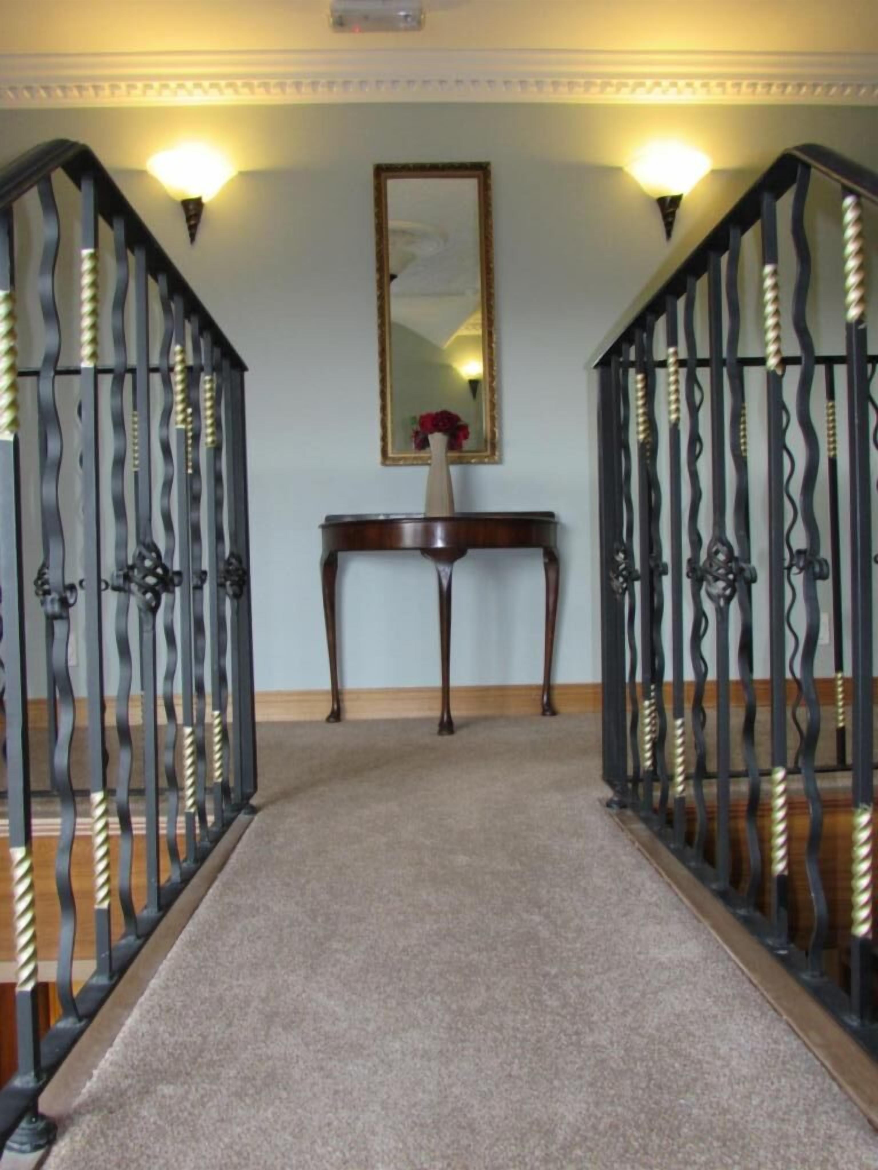 Un pasillo con suelo alfombrado, una consola de madera con un jarrón de flores y dos puertas de metal negro con detalles decorativos.