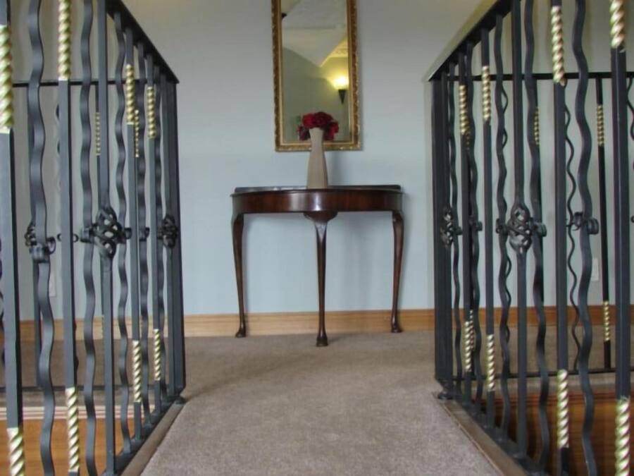 Un pasillo con suelo alfombrado, una consola de madera con un jarrón de flores y dos puertas de metal negro con detalles decorativos.