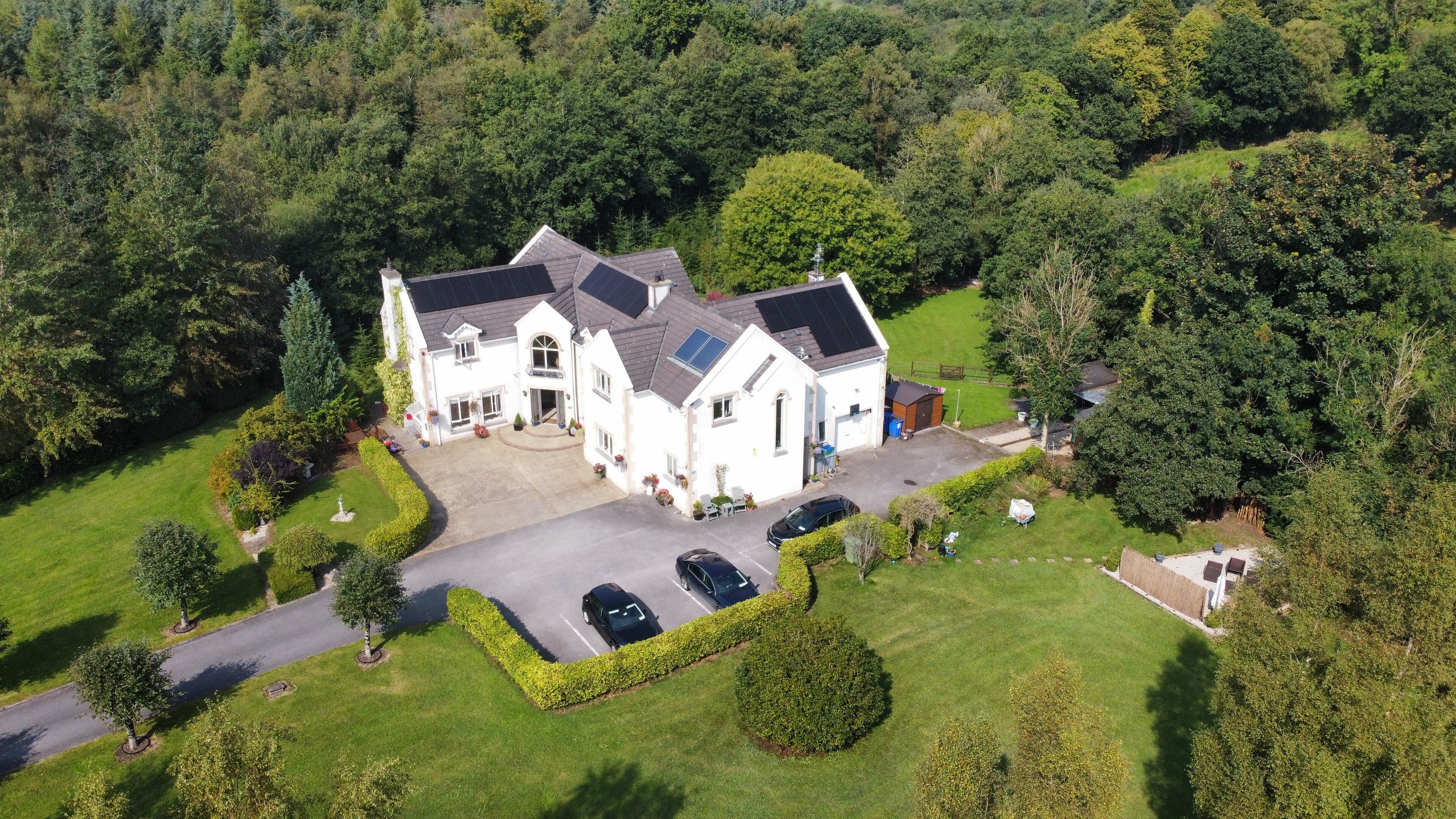 An aerial view of a large, white, two-story house with a driveway, parking area, and surrounding greenery.