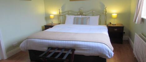 A neatly made bed with a headboard, two bedside tables with lamps, a wooden chair, a floral rug, and a framed picture on the wall.