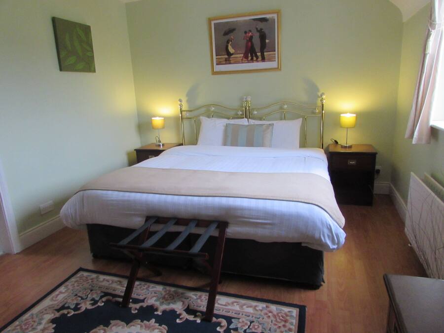 A neatly made bed with a headboard, two bedside tables with lamps, a wooden chair, a floral rug, and a framed picture on the wall.