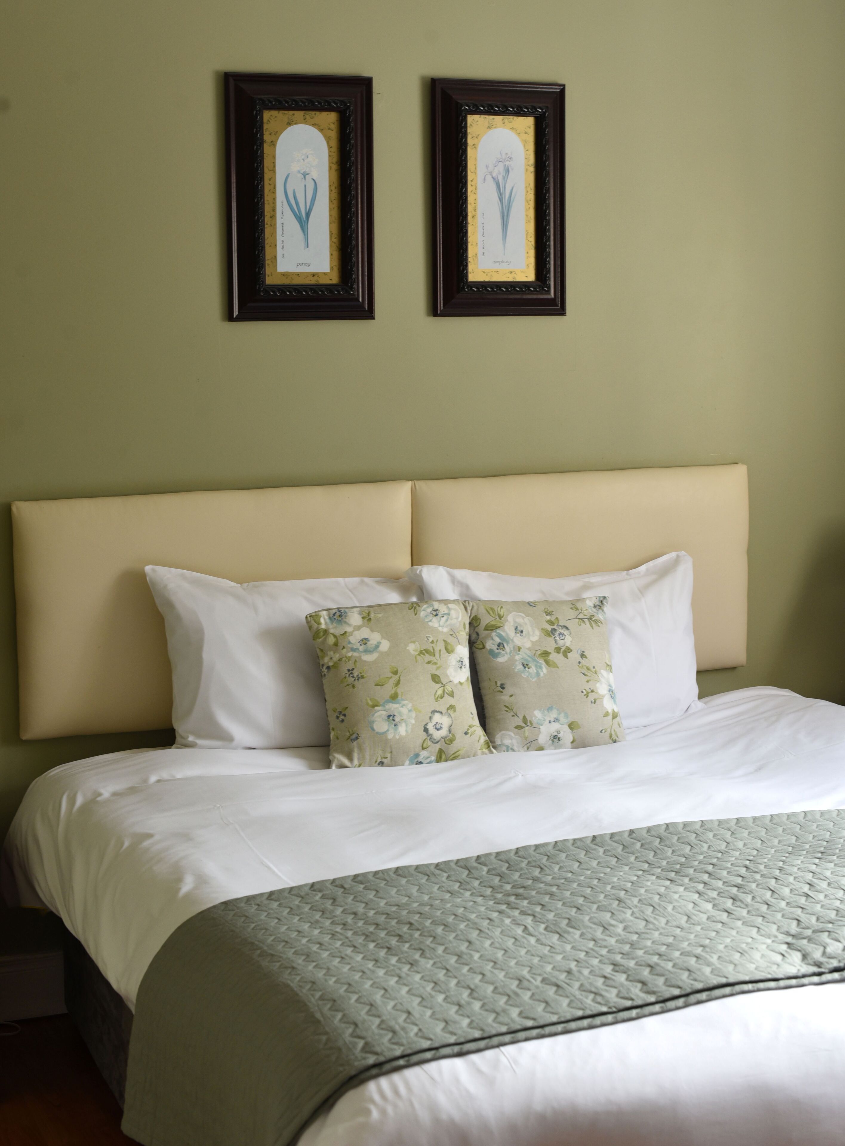 A neatly made bed with white and floral pillows, a beige headboard, and two framed botanical illustrations on the wall.