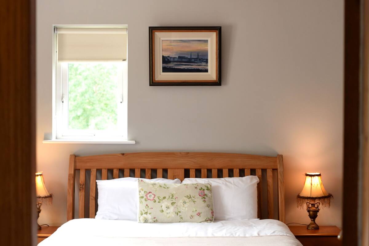 Un dormitorio con cama de madera, una ventana con vistas, un cuadro enmarcado en la pared y dos lámparas de noche.
