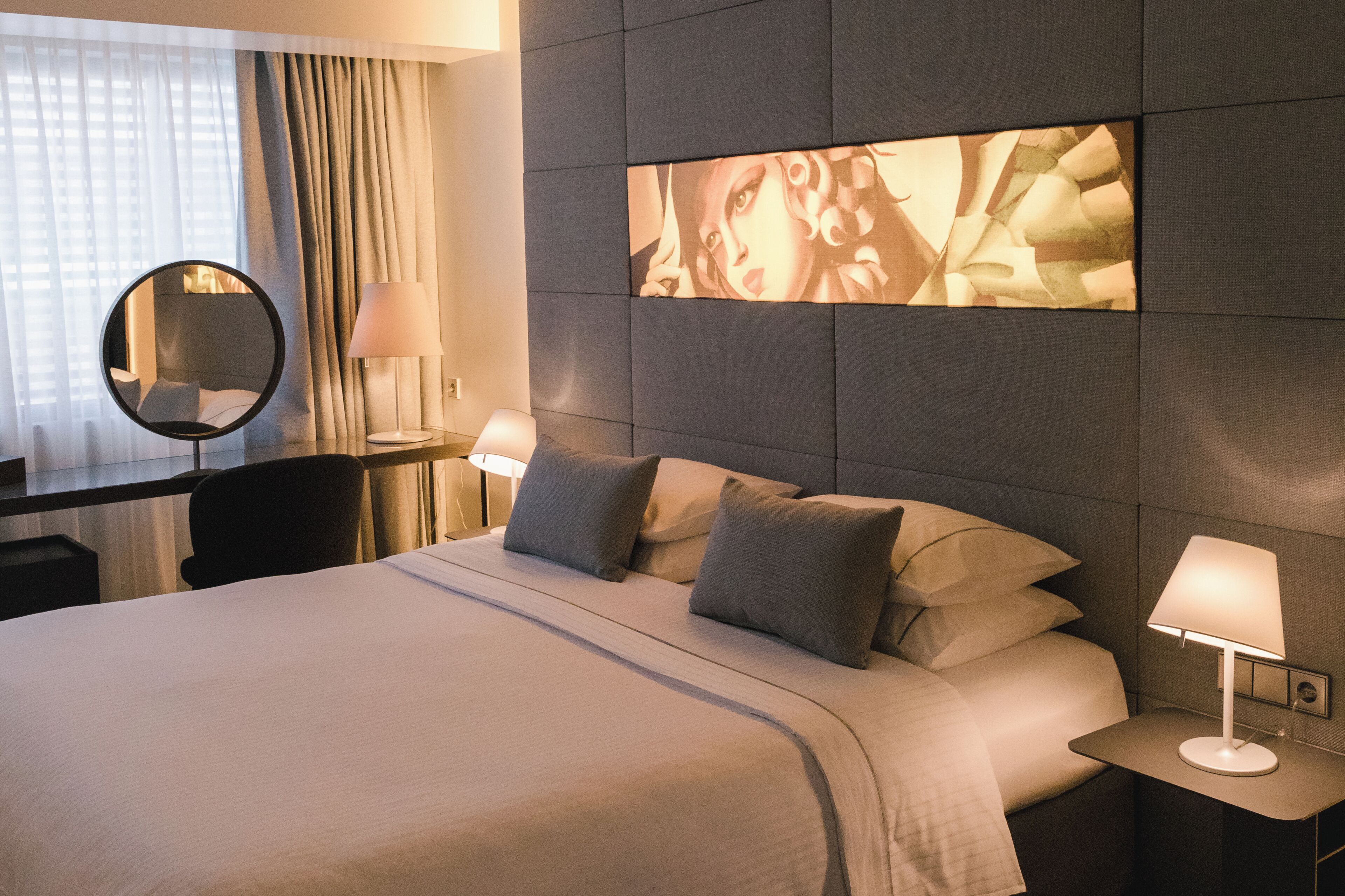 A hotel room with a large bed, a bedside table with a lamp, a desk with a lamp, a mirror, and a wall with a large artwork.