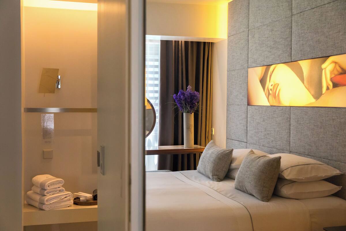 A modern hotel room with a neatly made bed, a bedside table with towels, a vase with purple flowers, and a large wall-mounted artwork.