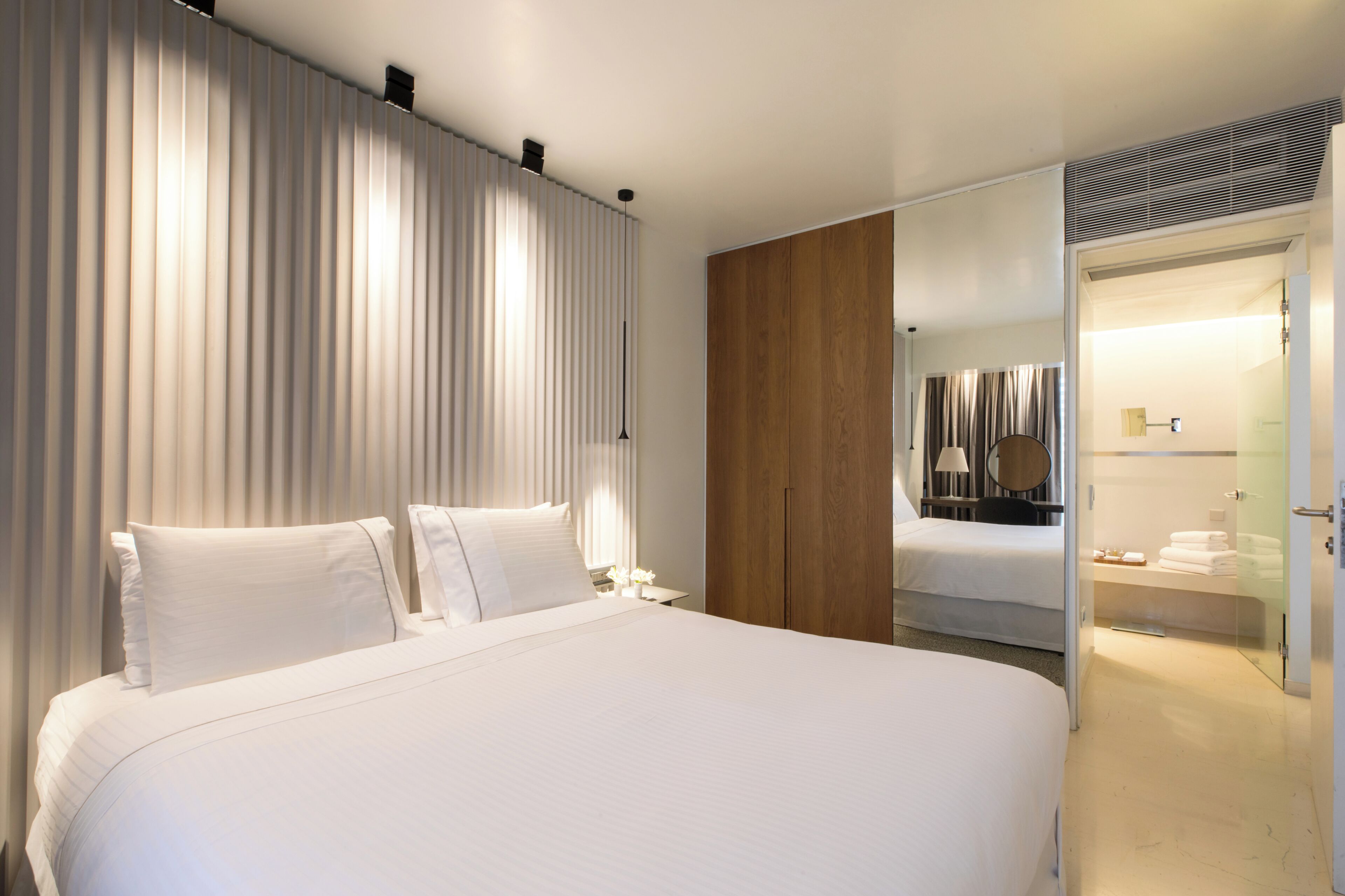 A modern hotel room with a large bed, a wooden wardrobe, a bathroom with a glass shower, and a bedside table with a lamp.