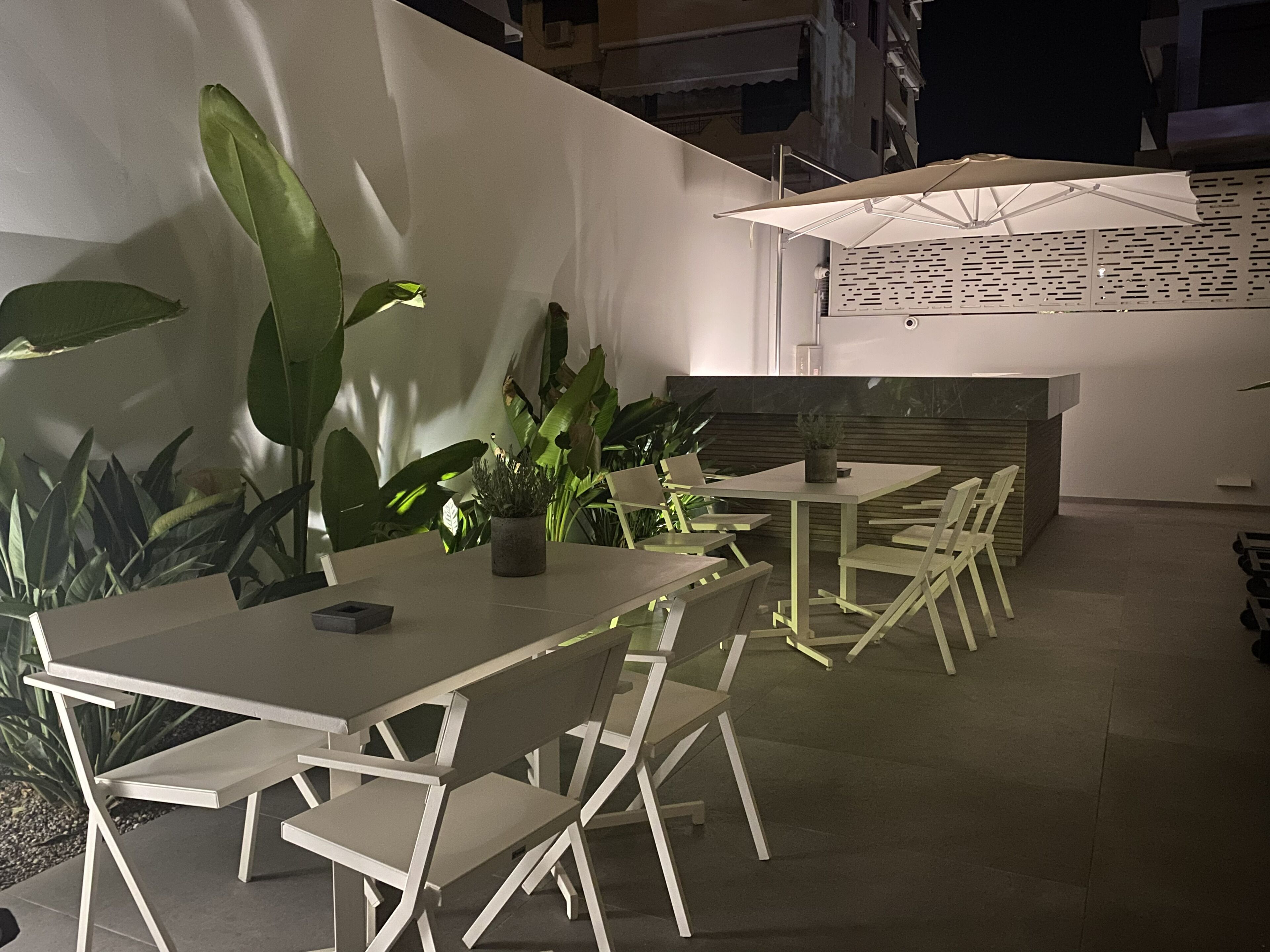 Un espacio al aire libre moderno con mesas y sillas blancas, rodeado de grandes plantas verdes.