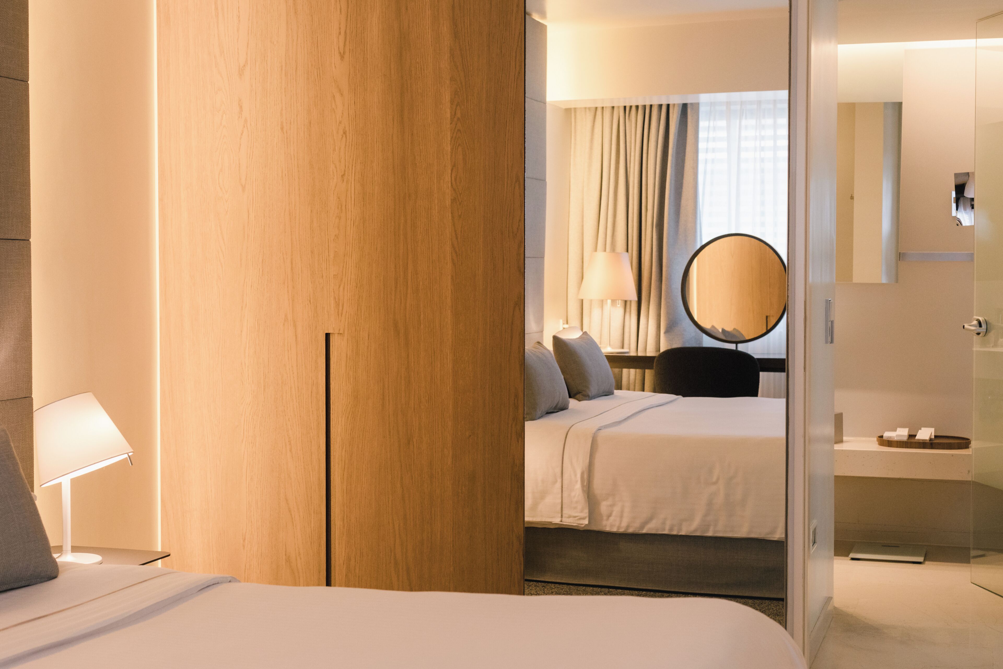 Een hotelkamer met een bed, nachtkastje, lamp, spiegel en een badkamer met een wastafel en handdoekhouder.