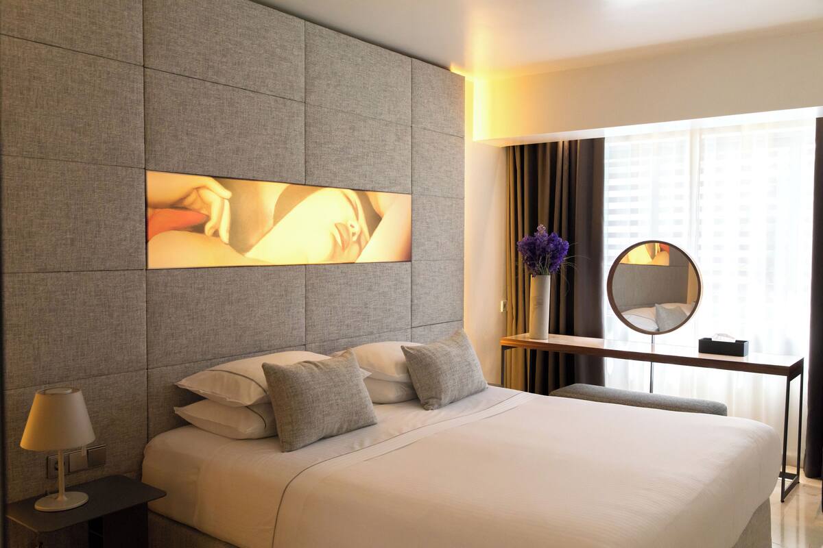 A modern hotel room with a large bed, a bedside table with a lamp, a mirror, and a view of the city through the window.