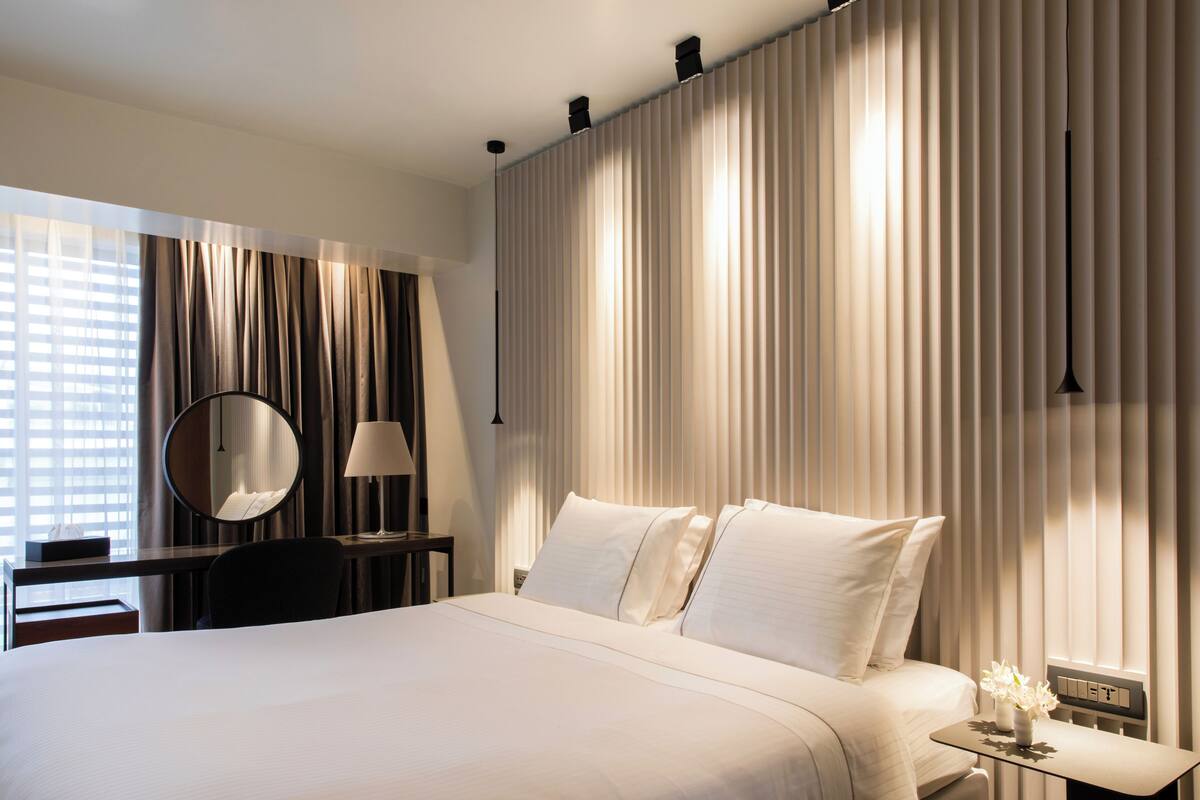 A hotel room with a large bed, a desk with a lamp, a mirror, and a nightstand with a vase of flowers.