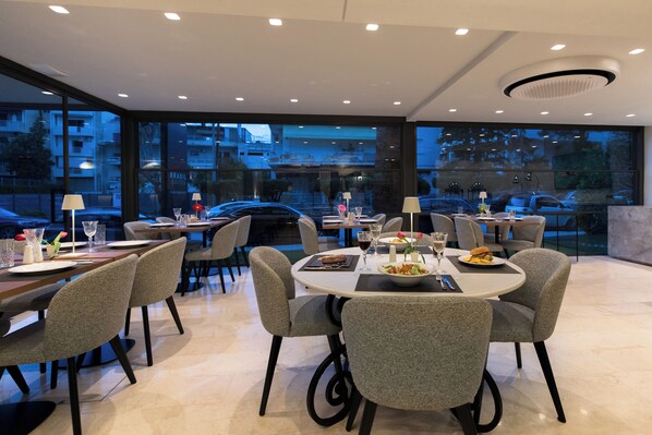A modern dining area with round tables and grey upholstered chairs, large windows offering a view of the cityscape, and a clean, contemporary design. - Brasil Suites Boutique Hotel (Glyfada)