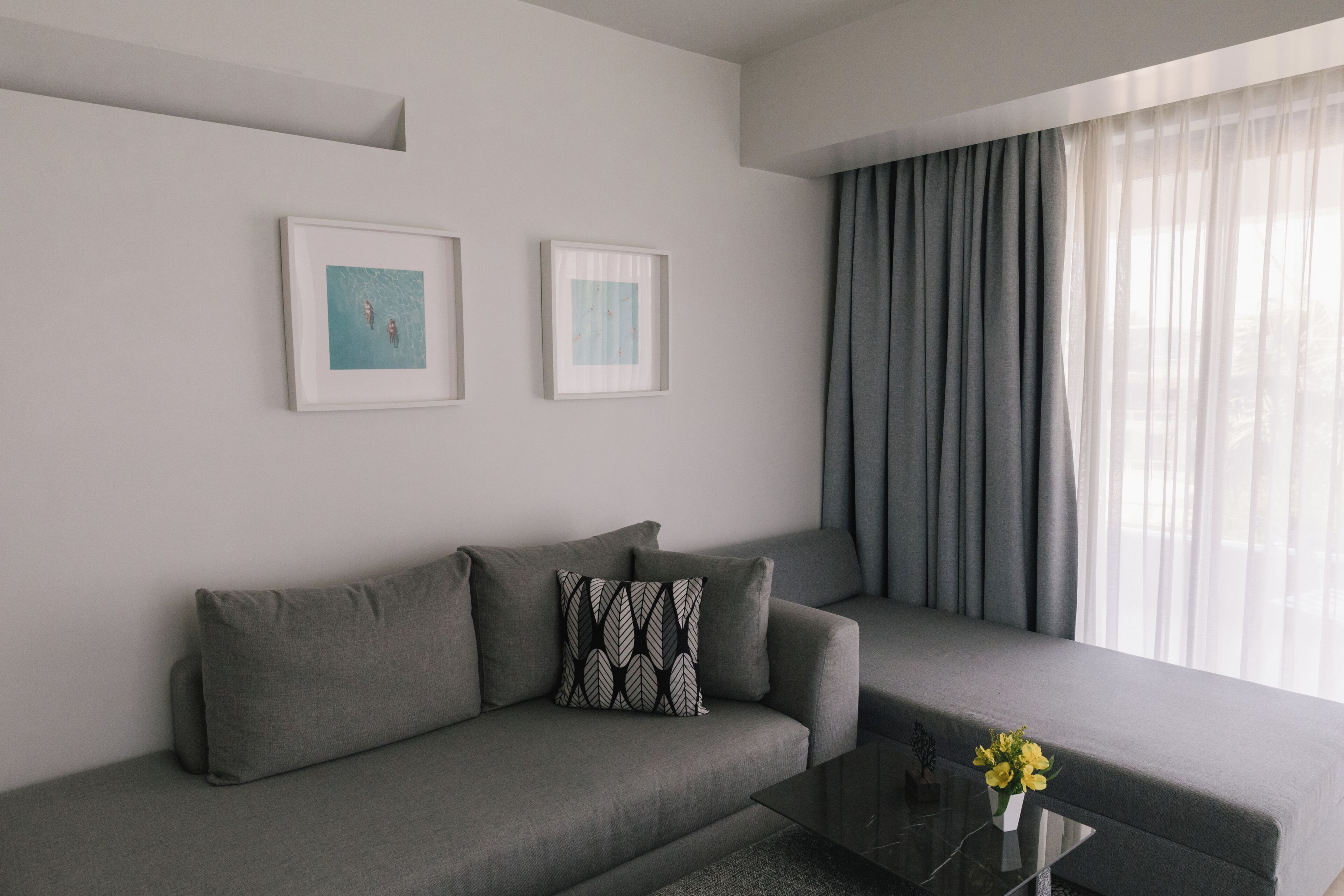 Een moderne woonkamer met een grijze bank, twee ingelijste kunstwerken, een glazen salontafel met een vaas gele bloemen, en een groot raam met grijze gordijnen.