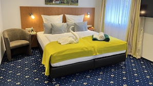 Habitación de hotel con una cama grande, cabecera de madera, mesita de noche con lámpara, una silla, un cuadro de un paisaje montañoso y un televisor.