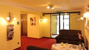 Un vestíbulo de hotel con alfombra roja, un sofá marrón, una mesa con un periódico y un revistero de pared.