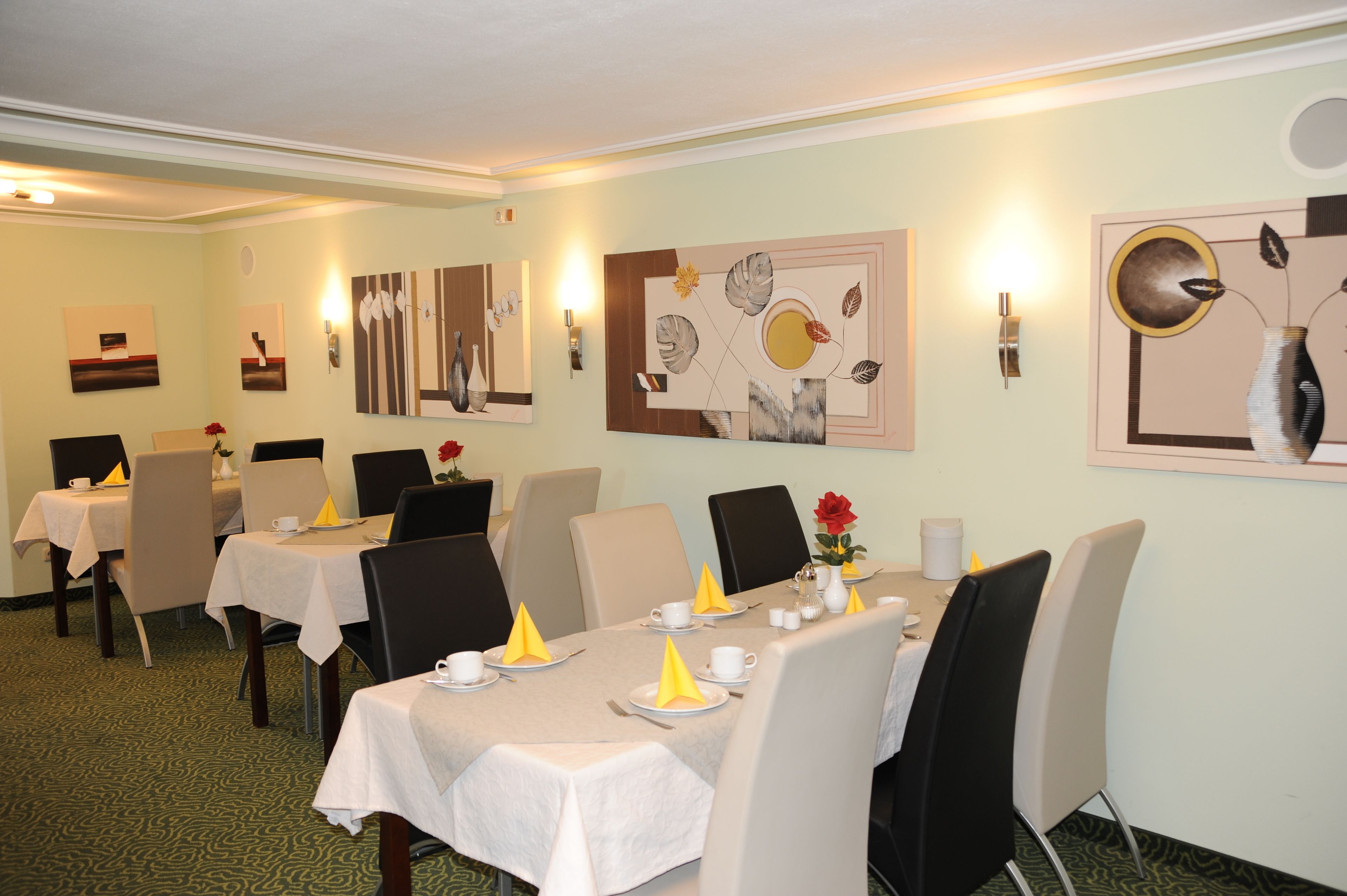 A dining area with tables set for four, each with white tablecloths, yellow napkins, and red flowers. The walls are adorned with abstract art.