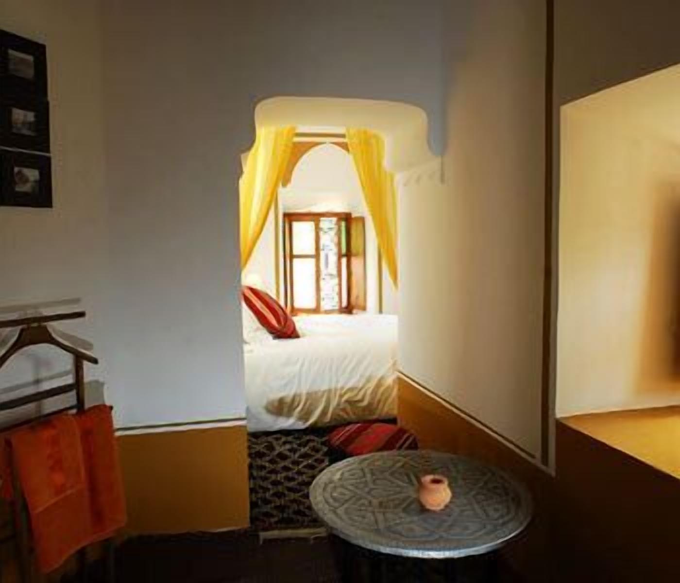 A narrow hallway leading to a bedroom with a bed, a small table with a vase, and a window with yellow curtains.
