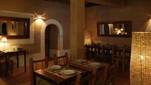 A warmly lit dining area with wooden tables and chairs, a large arched doorway, and a mirror on the wall. - Kasbah Dar Essalam (Skoura)