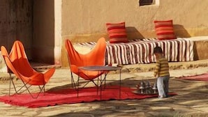 A room with a sofa, two orange chairs, a small table, and a child standing near a metal object. - Kasbah Dar Essalam (Skoura)