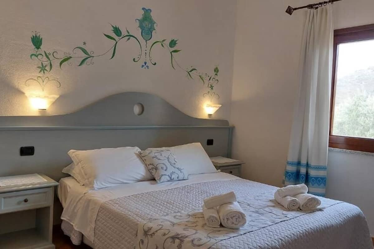 Ein ordentlich bezogenes Bett mit weißer Bettwäsche und dekorativen Kissen, ein Nachttisch mit Lampe und Wandkunst.