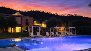 Une piscine avec des chaises longues, un bâtiment doté d’un toit en tuiles et un coucher de soleil en arrière-plan.