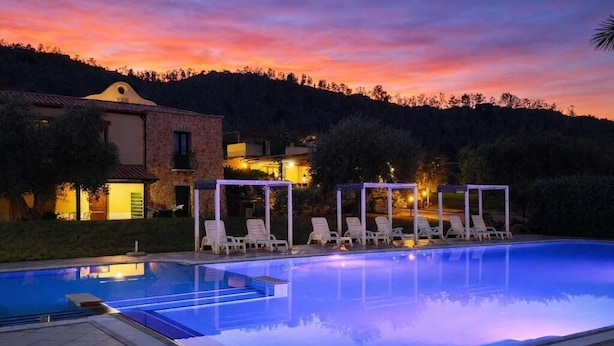 Una piscina con sillones de descanso, un edificio con techo de tejas y una puesta de sol al fondo.