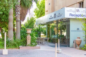 Zona de recepción de hotel con fachada de vidrio, asientos exteriores y plantas tropicales.