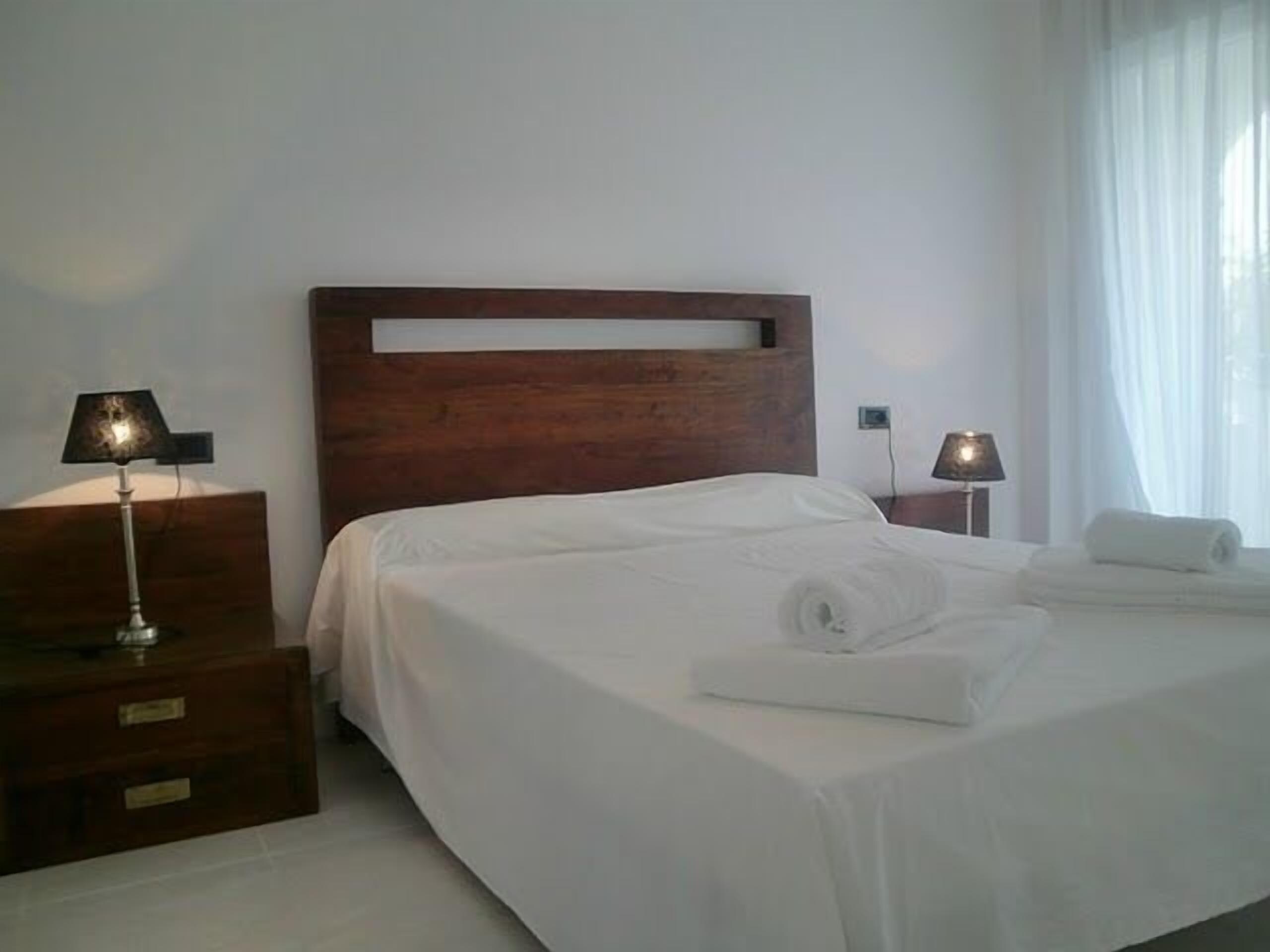 Een net opgemaakt bed met witte lakens en een opgerolde handdoek, een houten hoofdbord en twee bedlampen op houten nachtkastjes.