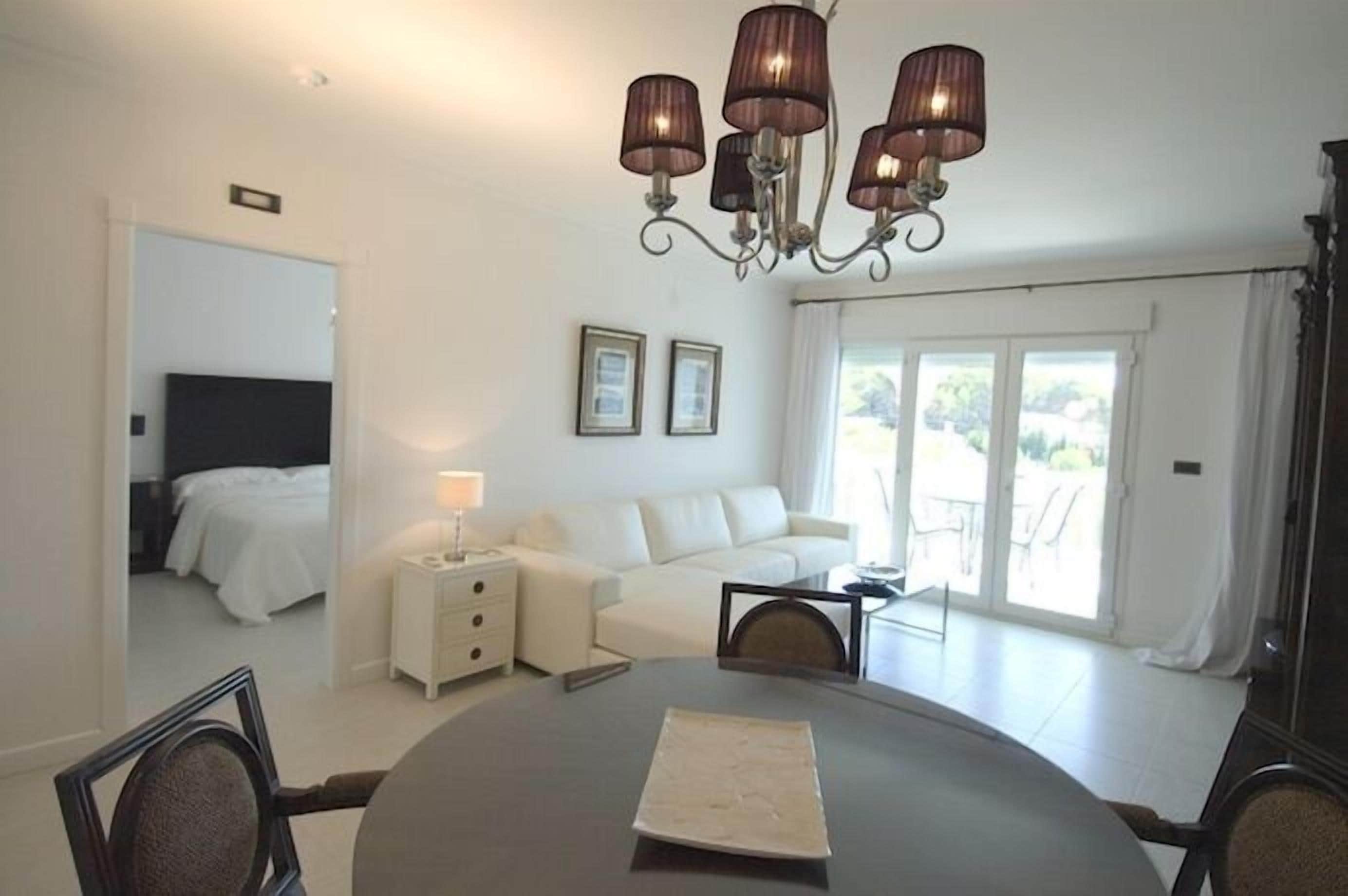 A modern hotel room with a dining table, a sofa, a chandelier, and a view of the outdoors through the sliding glass doors.