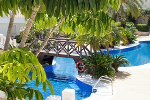 A swimming pool with a bridge, surrounded by palm trees and other greenery. - Aparthotel Residencia & Spa Parques Casablanca (Benissa)