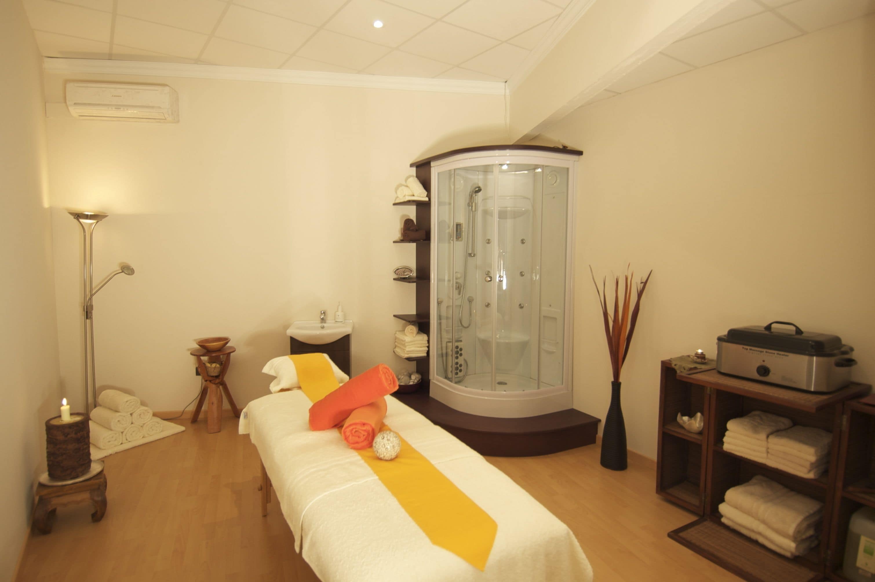 Une salle de spa équipée d’une table de massage, d’une douche à vapeur à parois de verre, d’un petit lavabo et d’une étagère avec des serviettes.