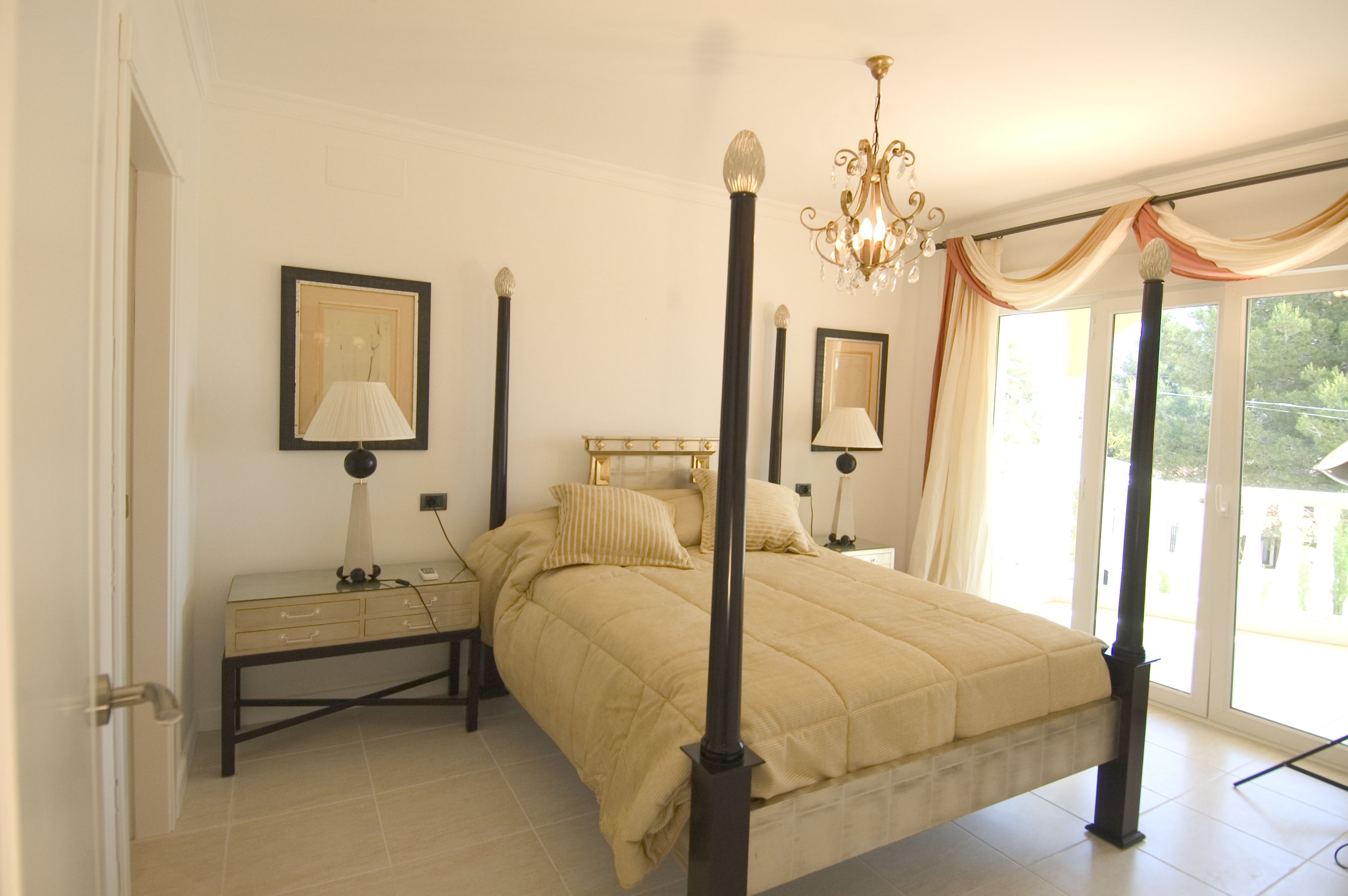 A bedroom with a four-poster bed, a nightstand, a lamp, and a view of the outdoors through a large window.