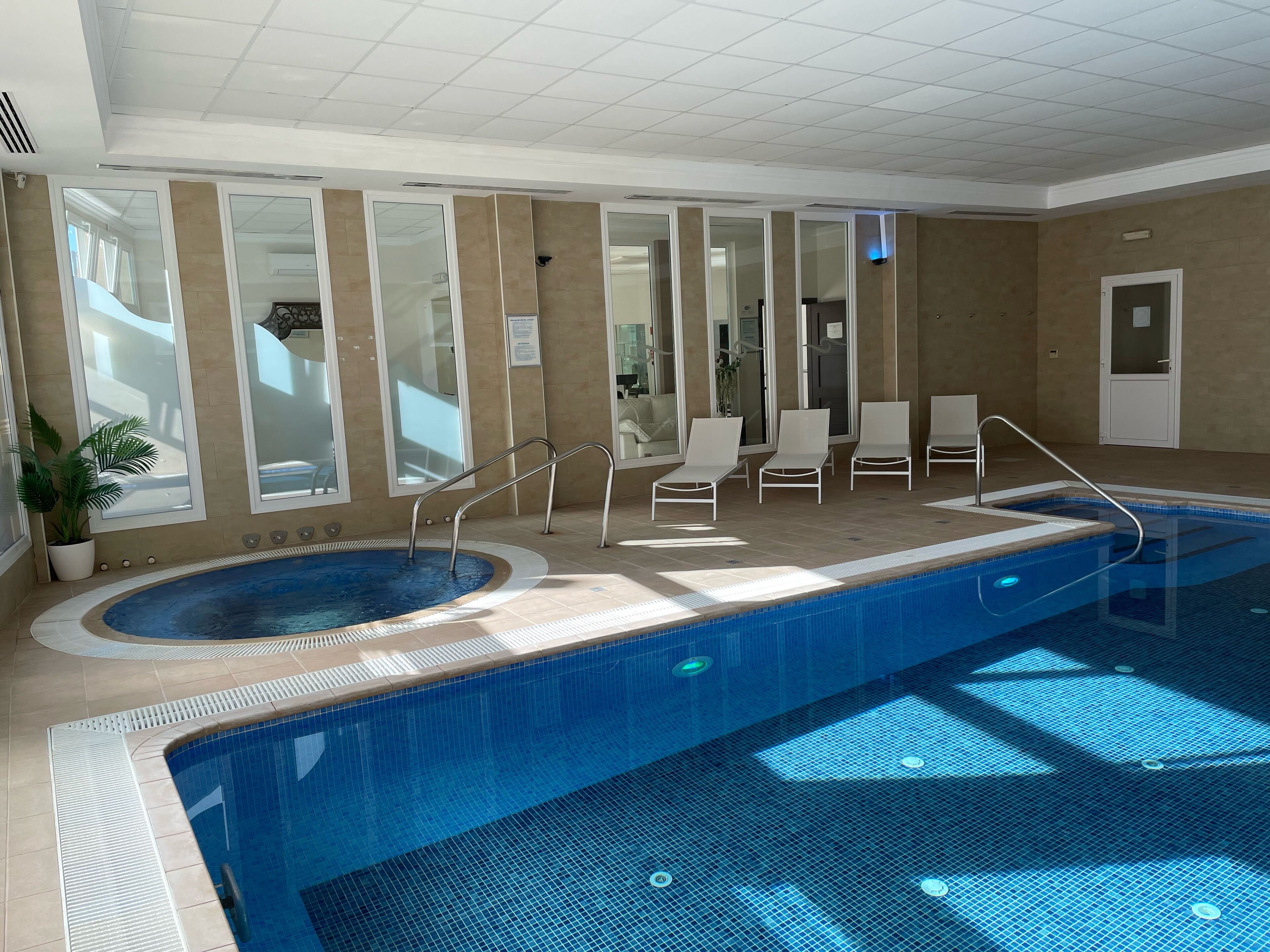 An indoor swimming pool with a hot tub, sunlit by large windows, and equipped with lounge chairs and a safety railing.