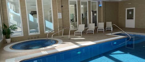 An indoor swimming pool with a hot tub, sunlit by large windows, and equipped with lounge chairs and a safety railing.
