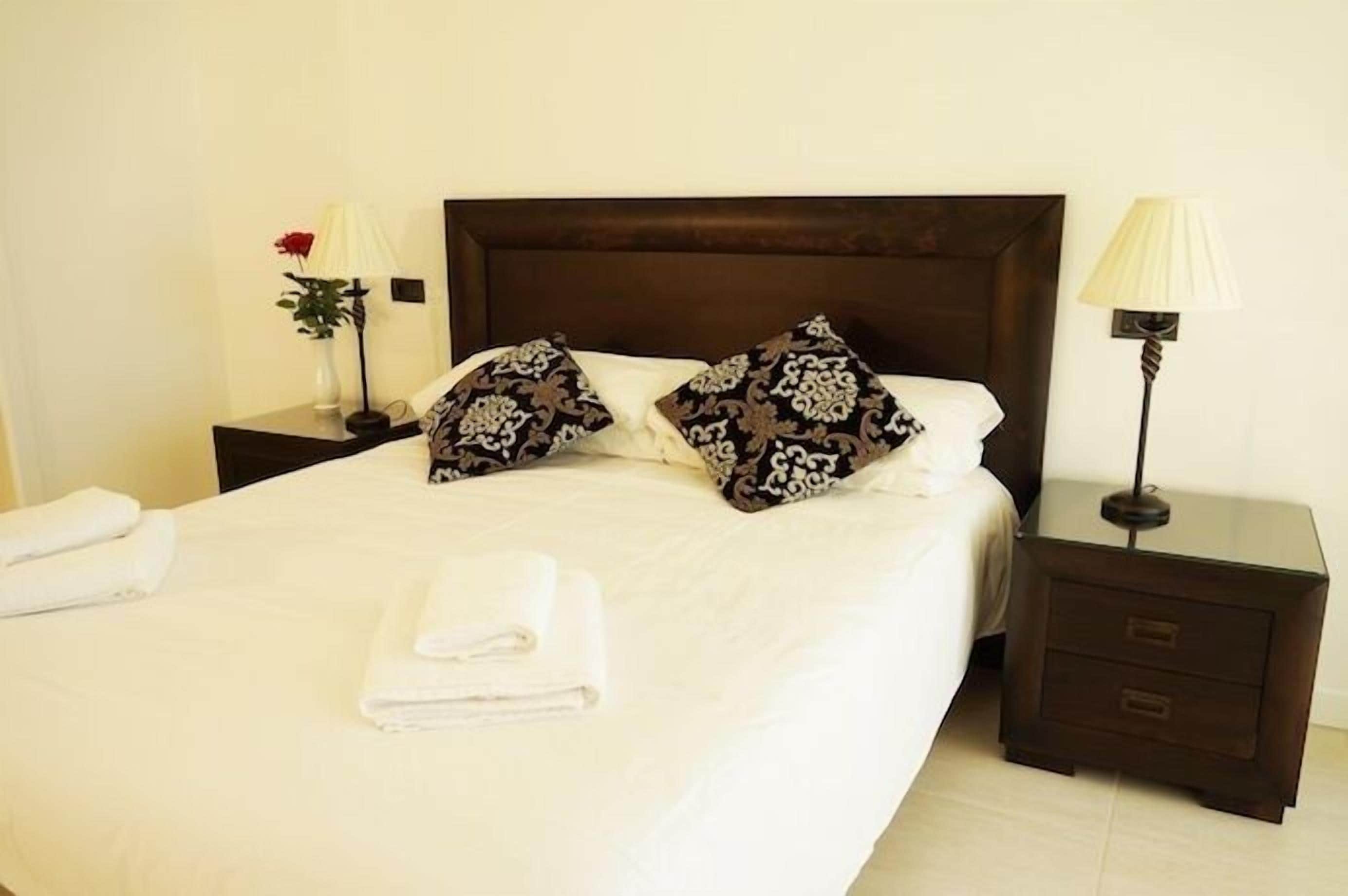 A neatly made bed with white linens and two patterned pillows, a bedside table with a lamp, and a vase with a red flower.