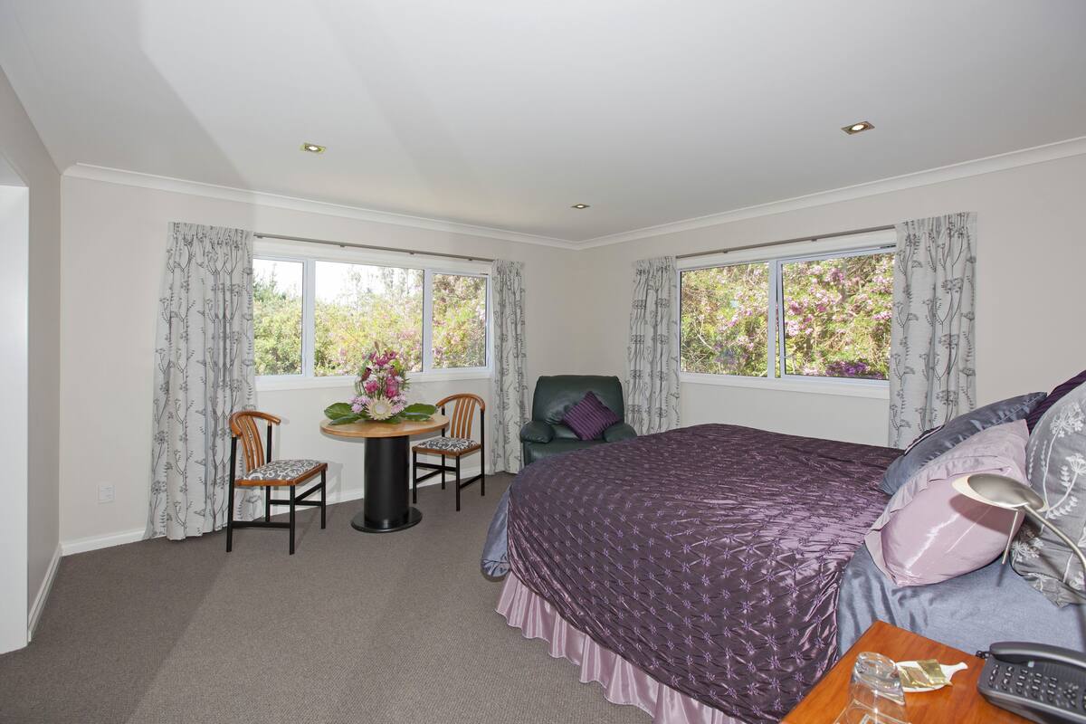A bedroom with a large bed, two windows with curtains, a chair, a small table with a vase, and a desk with a telephone.