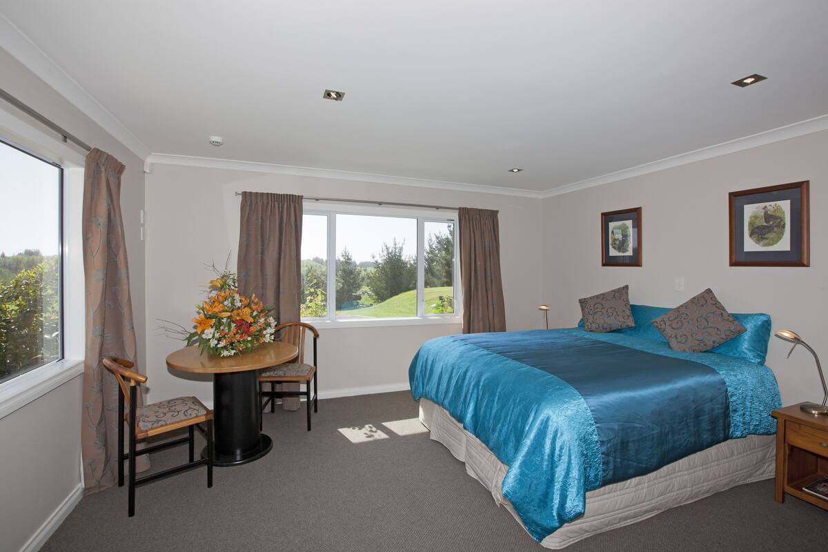 A bedroom with a large bed, a desk with a chair, a window with curtains, and framed pictures on the wall.