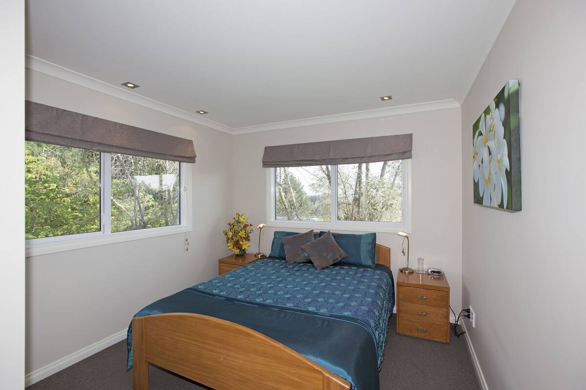 A bedroom with a bed, two bedside tables, a nightstand, a window with a view of trees, and a wall-mounted picture.