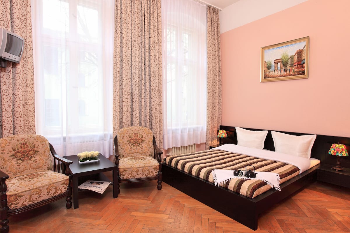 Quarto com uma cama, duas cadeiras, uma mesinha, um abajur e um quadro na parede.
