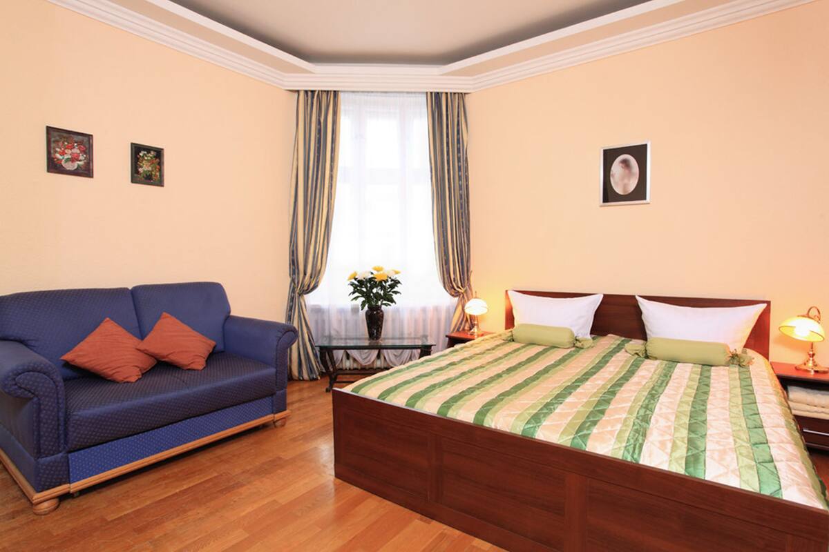 Quarto de hotel com piso de madeira, um sofá azul com almofadas vermelhas, uma cama com colcha listrada, uma mesa de cabeceira com abajur e uma janela com cortinas.