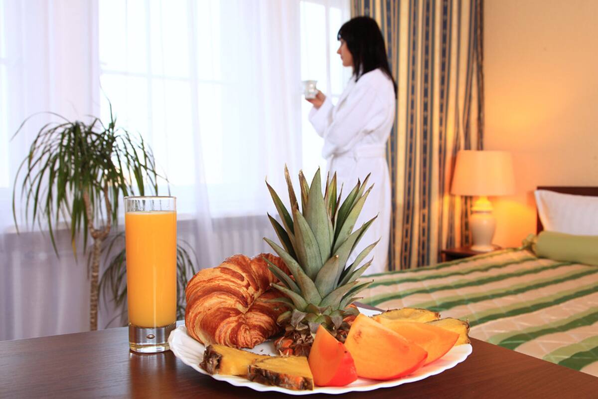 Mesa de café da manhã com uma travessa de frutas e um croissant, um copo de suco de laranja, e uma pessoa ao fundo.