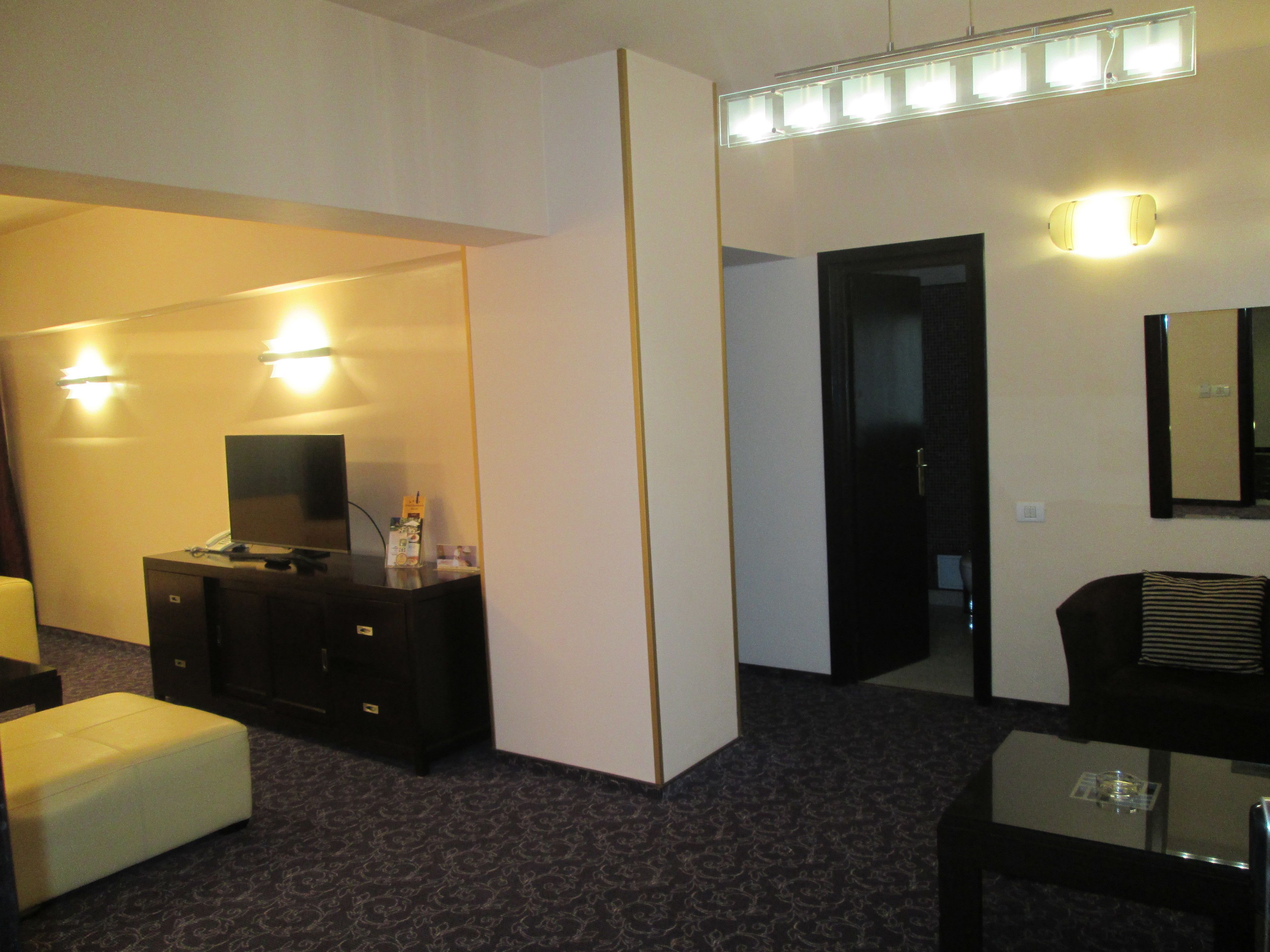 Ein Hotelzimmer mit einem Flachbildfernseher auf einer dunklen Holzkommode, einem weißen Hocker, einem Glastisch und einem Spiegel an der Wand.