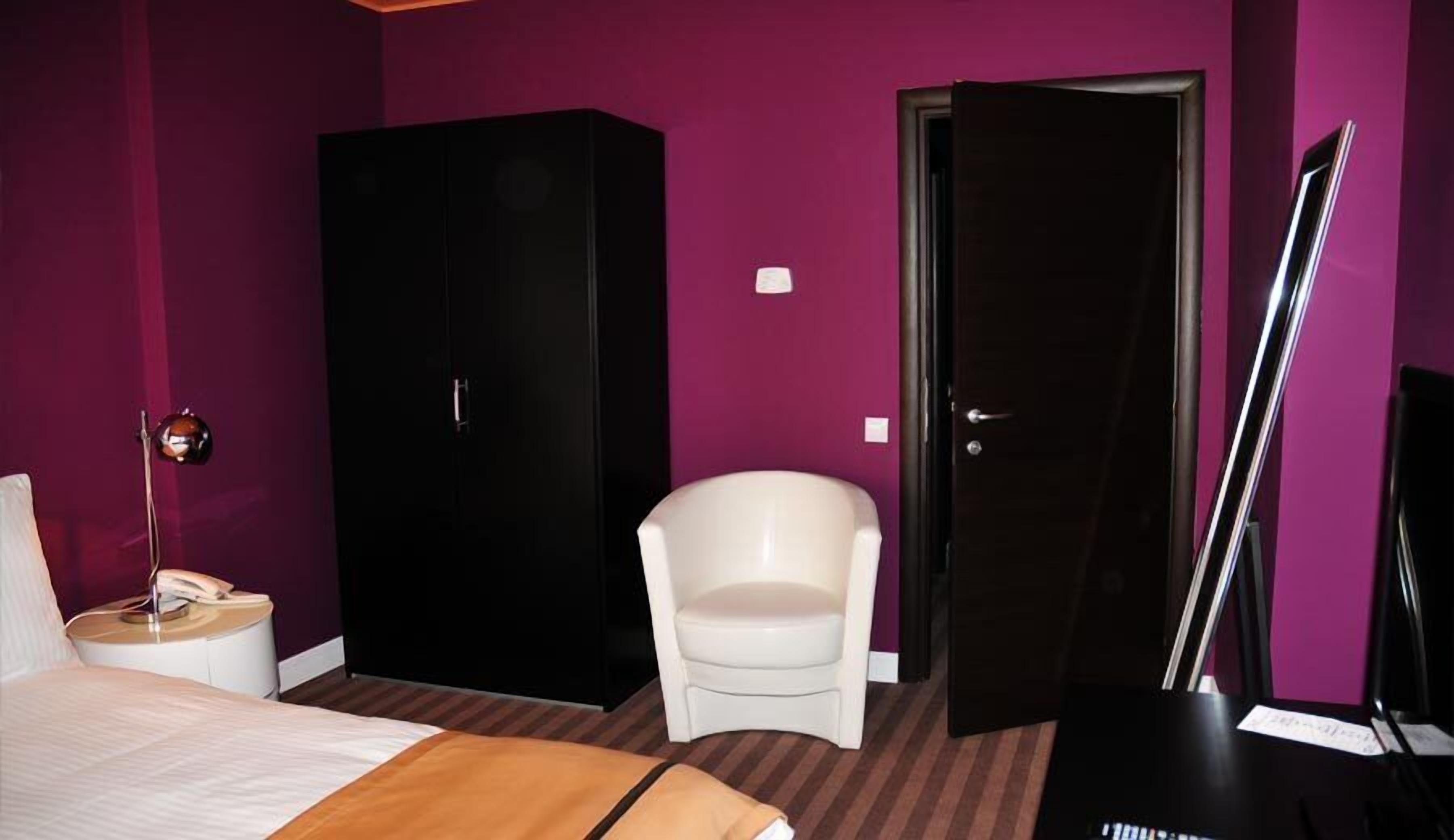 A hotel room with a bed, a white chair, a black wardrobe, and a door.