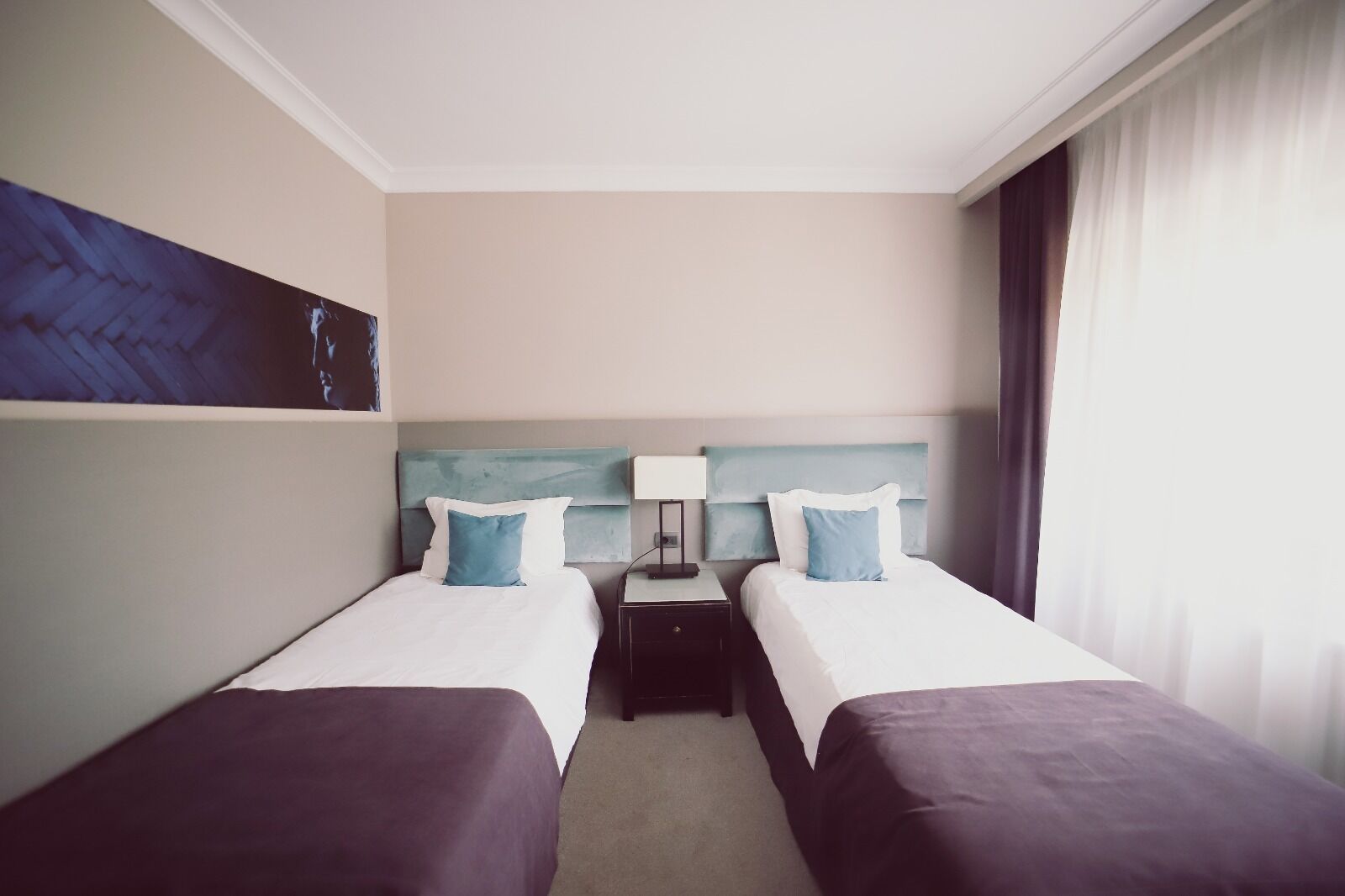 A hotel room with two beds, a nightstand, a lamp, and a painting on the wall.