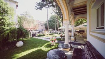 An outdoor seating area with tables and chairs, surrounded by greenery and a building with a yellow facade.