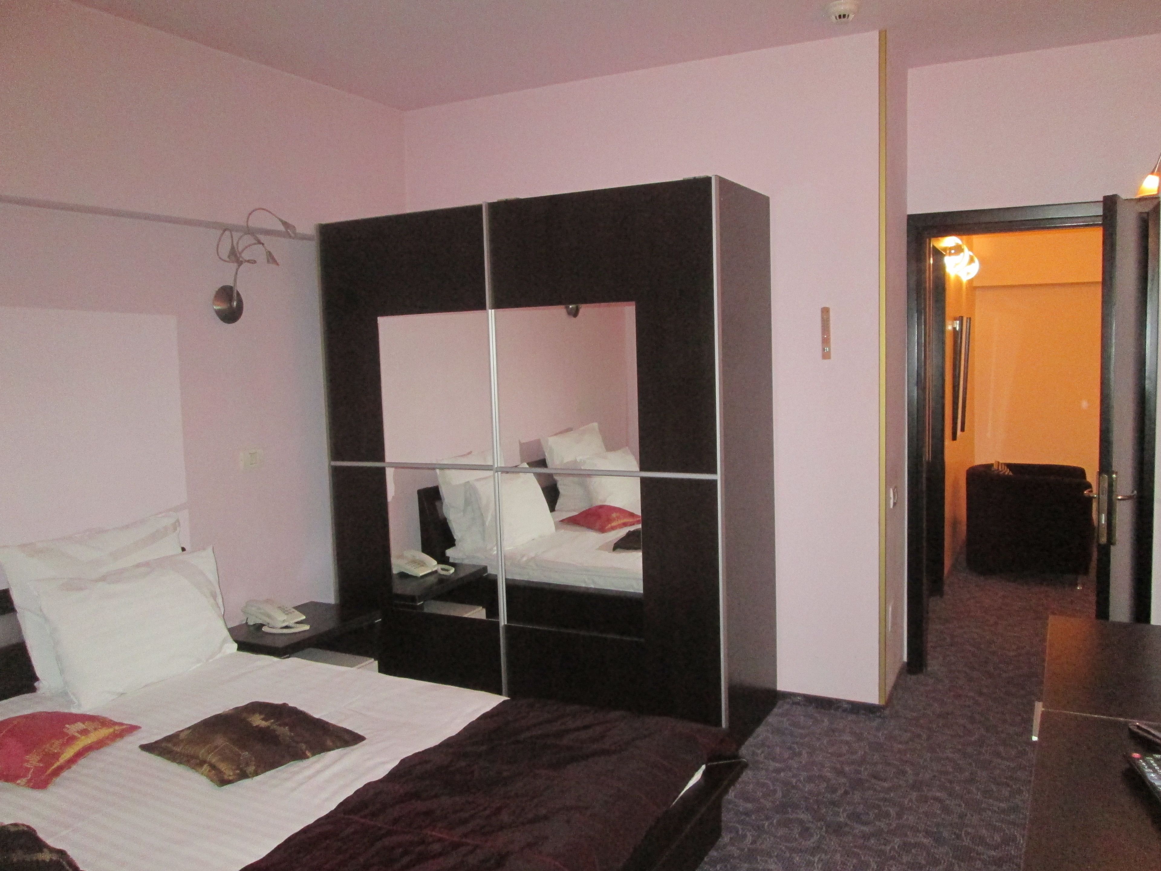 Una habitación de hotel con una cama grande, un armario, una mesita de noche con teléfono y una puerta que da a un pasillo.