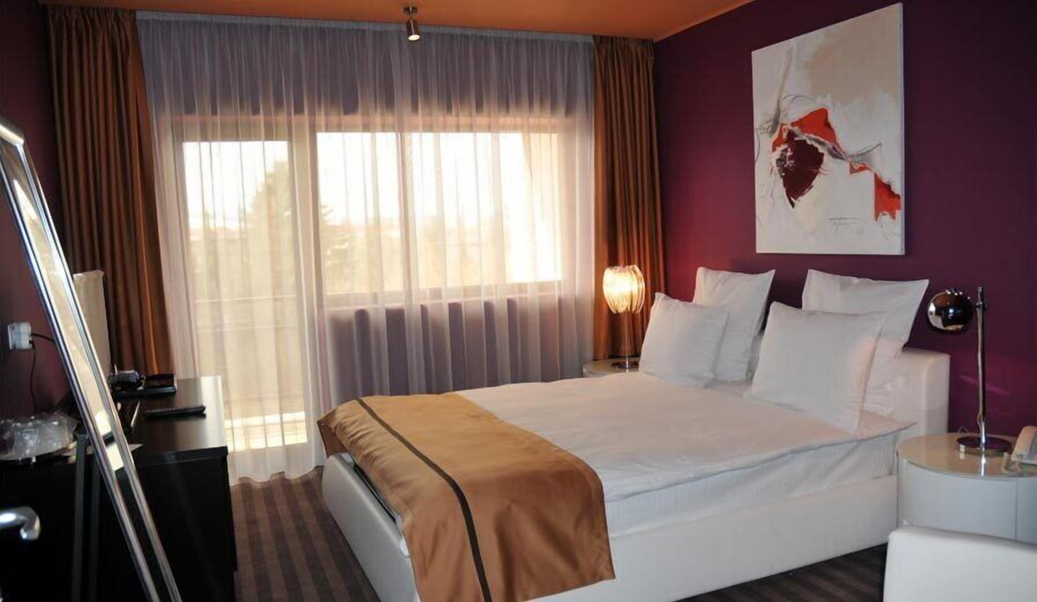 Una habitación de hotel con una cama grande, un escritorio, una silla, una lámpara y un cuadro en la pared.