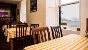 A dining area with checkered tablecloths and wooden chairs, a window with a view of the sea, and a framed picture on the wall.